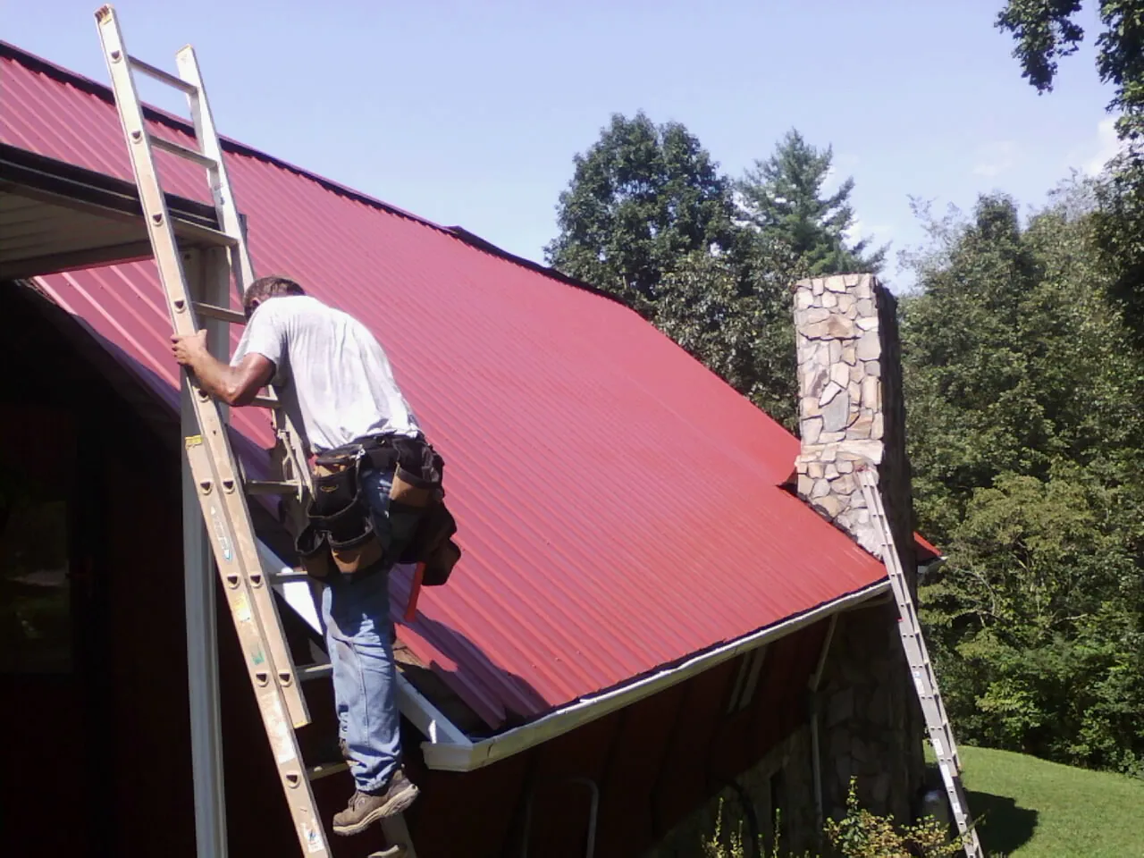 Roofer on a ladder beside a red metal roof and stone chimney — Soffit & Fascia Repair by Roofing Brothers in Scituate