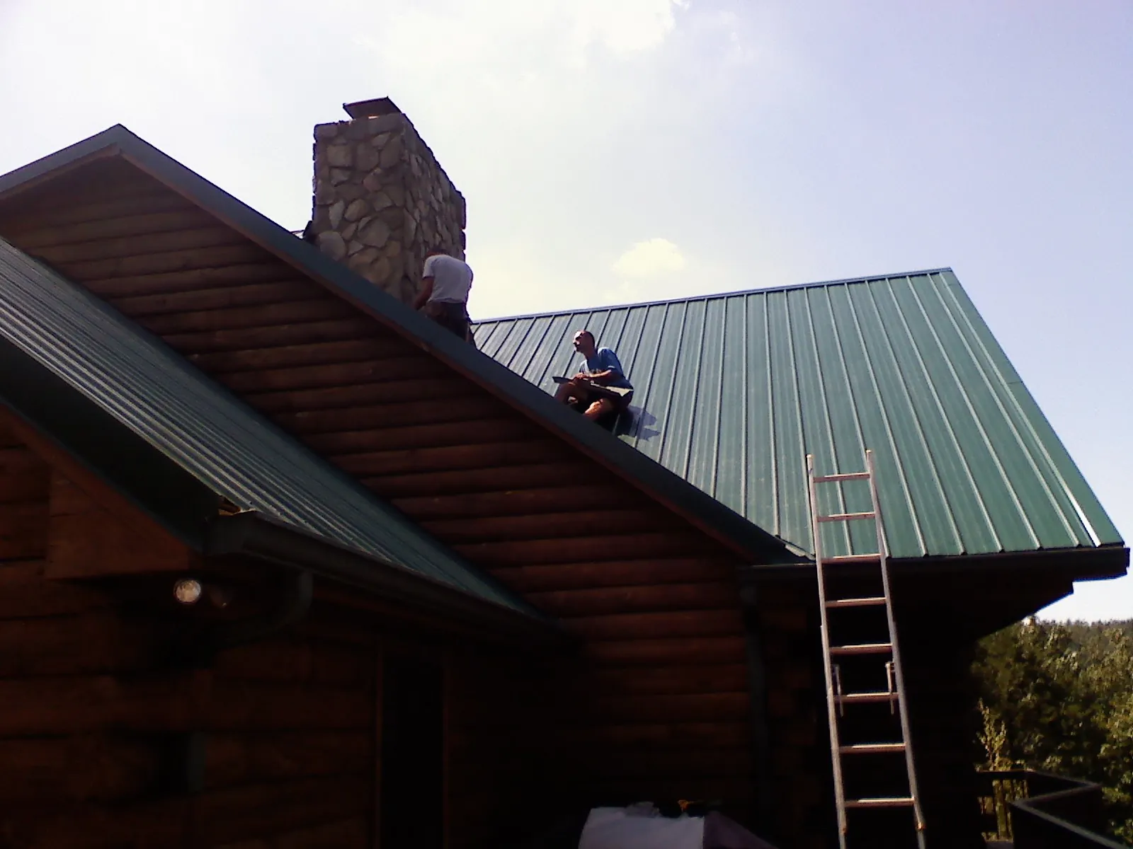 Roofers installing green metal panels beside a stone chimney on a log home in Scituate